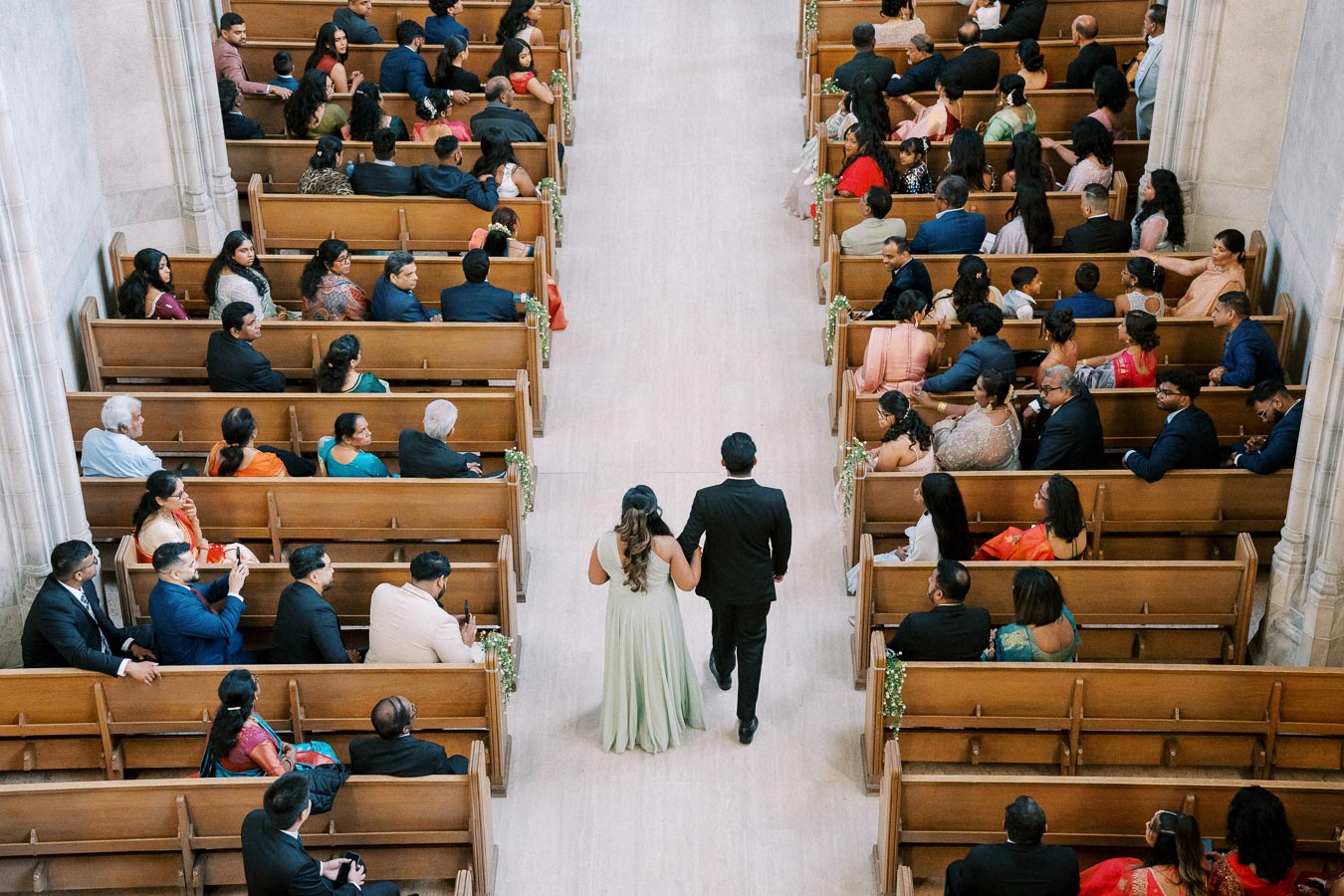 Overhead view of a wedding ceremony in a church with a couple walking down the aisle, surrounded by seated guests in formal attire.