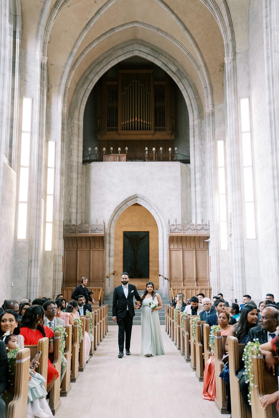 A couple walking down the aisle in a beautifully decorated church with tall ceilings and guests watching from the pews.