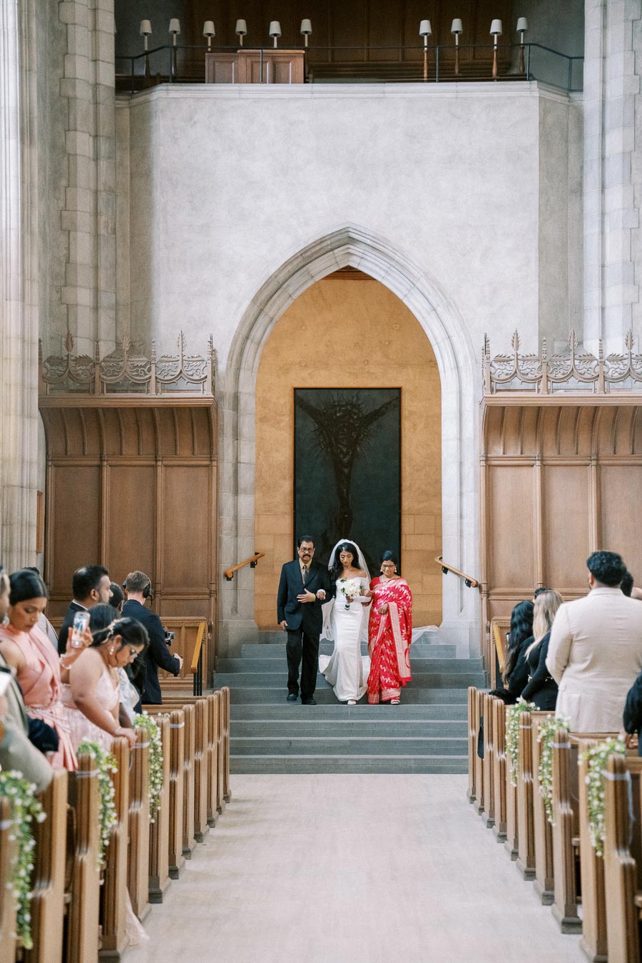 Bride walking down the aisle with parents in a grand cathedral; wedding guests seated in beautifully decorated pews.