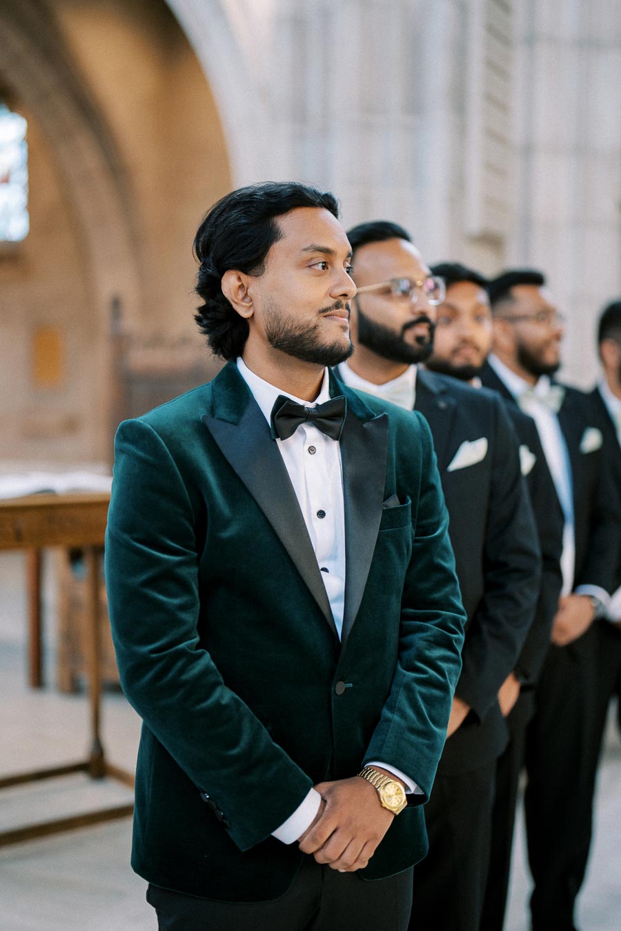 A groom in a velvet tuxedo jacket and bow tie stands confidently with his groomsmen in a formal setting.
