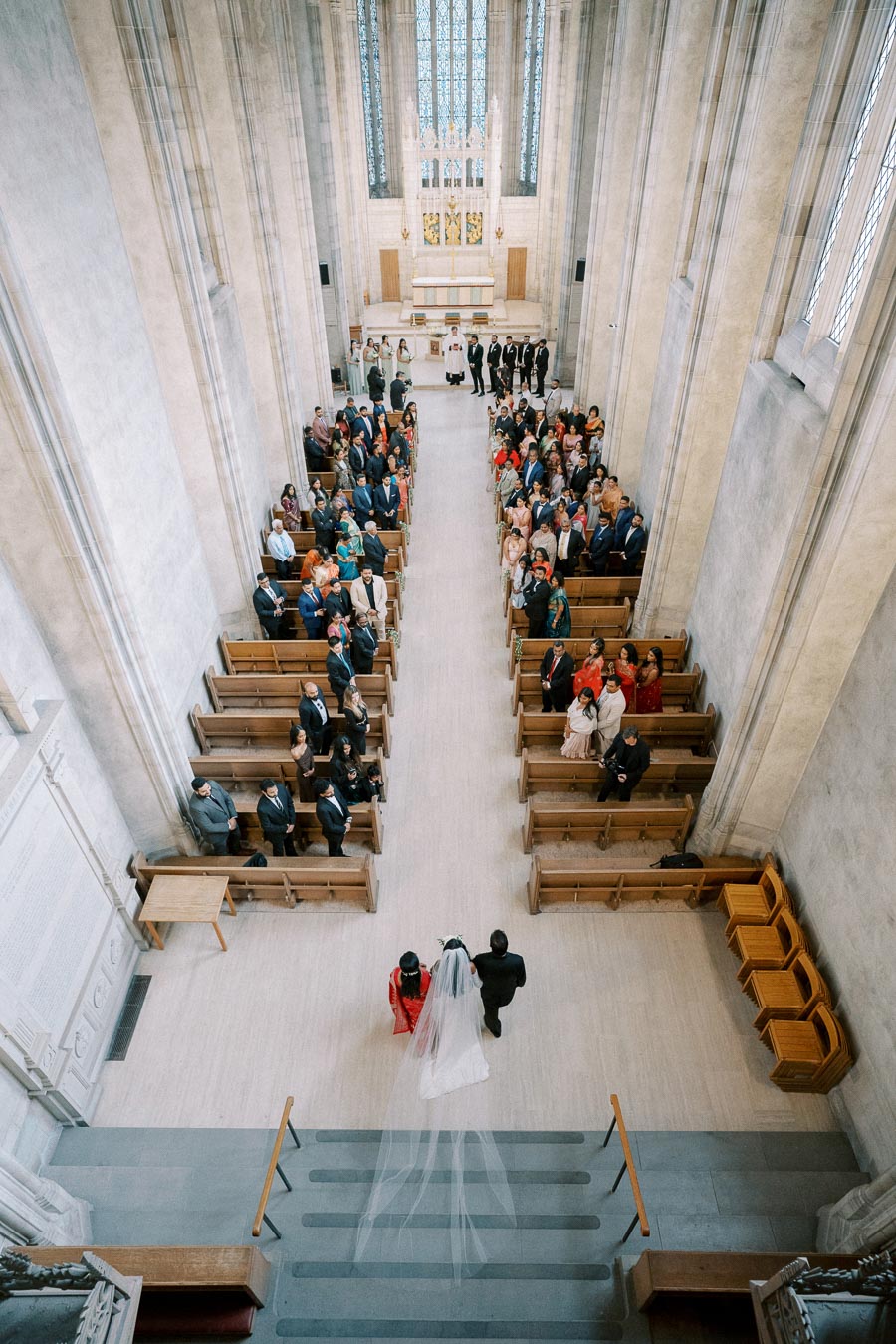 Wide-angle view of a wedding ceremony in a grand church, showcasing the bride and groom walking down the aisle lined with seated guests, capturing the architecture, stained glass windows, and elegant atmosphere.