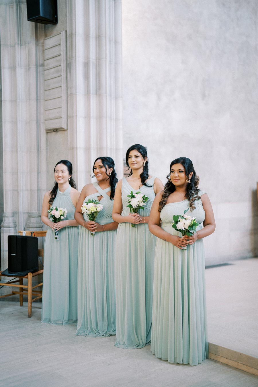 Bridesmaids in pastel blue dresses standing in a church setting, holding white floral bouquets, during a wedding ceremony.