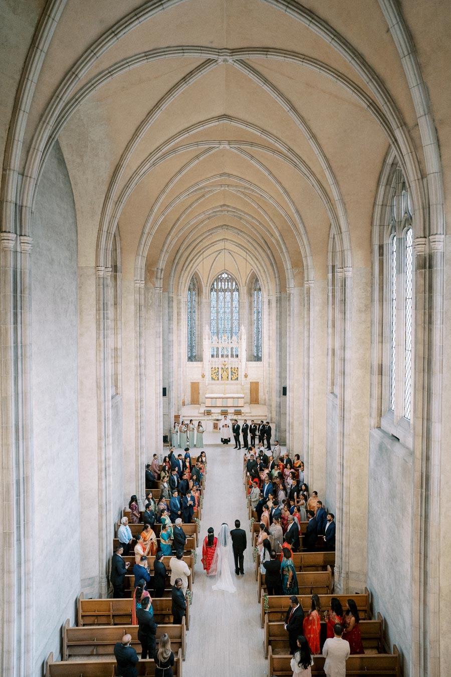 Aerial view of a wedding ceremony inside a grand cathedral with vaulted ceilings, featuring a bride and groom walking down the aisle surrounded by seated guests.