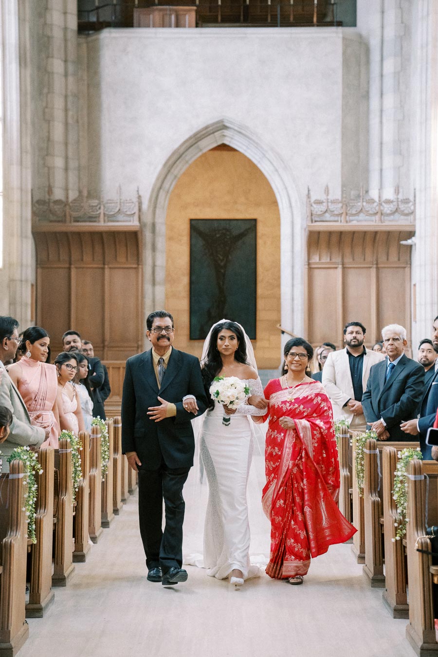 Bride walking down the aisle with her parents in an elegant church, surrounded by seated guests looking on during a wedding ceremony.