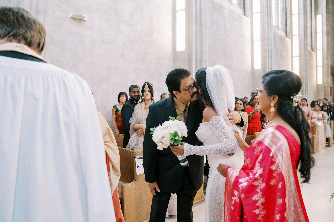 Father and bride embracing during a wedding ceremony in a church, with guests watching in the background. The bride is holding a bouquet of white flowers and wearing a lace gown and veil.
