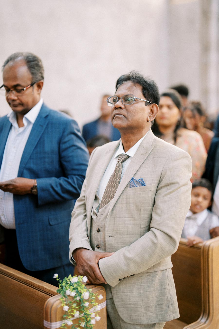 A man in a light beige suit and patterned tie stands attentively during a formal event, surrounded by other attendees seated in wooden pews.