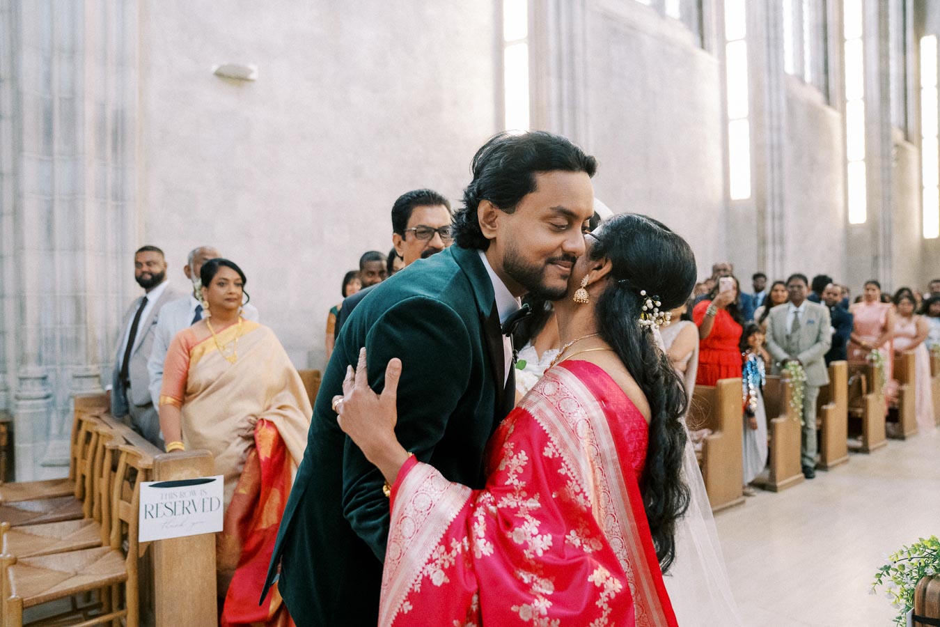 A couple embracing during a wedding ceremony in a church filled with guests, with the woman wearing a red sari and the man in a suit, surrounded by an attentive audience dressed in formal attire.