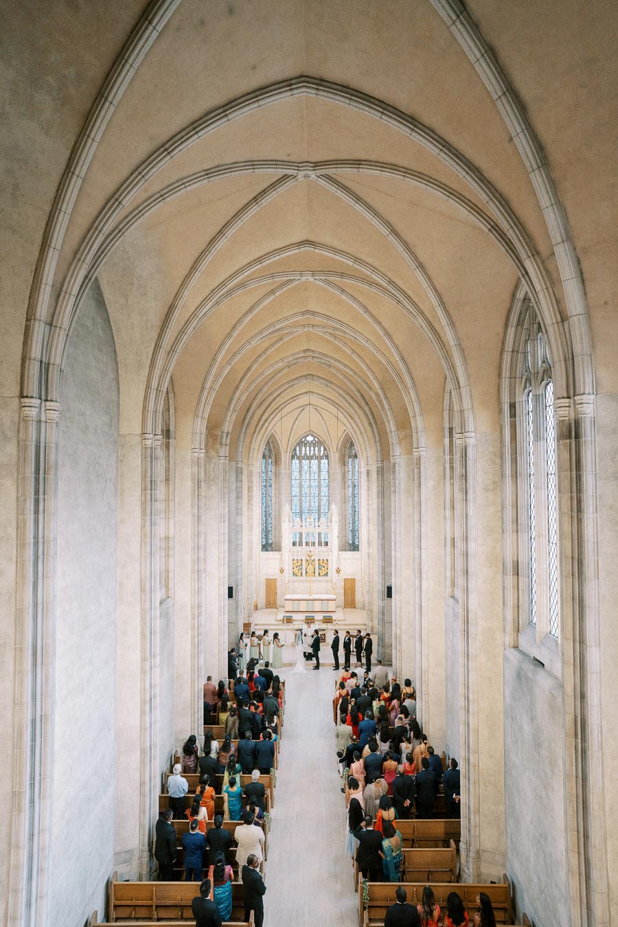 A wedding ceremony taking place in a large, ornate cathedral with guests seated in wooden pews, intricate vaulted ceilings, and a stained-glass window at the altar.