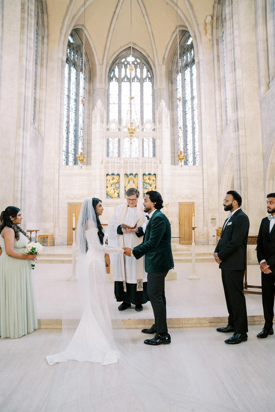 A bride in a white wedding dress and veil holds hands with the groom in a dark suit during their marriage ceremony at a grand cathedral. The officiant stands between them holding a book, while bridesmaids and groomsmen look on in the background. The cathedral features tall stained glass windows and ornate architecture.