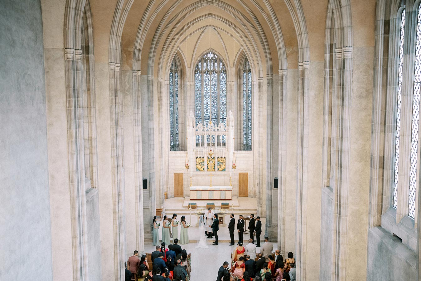 A serene wedding ceremony taking place in a grand, ornate church with vaulted ceilings, intricate architectural details, and large stained glass windows illuminating the altar. The bride and groom stand at the altar with the wedding party, surrounded by seated guests in formal attire.