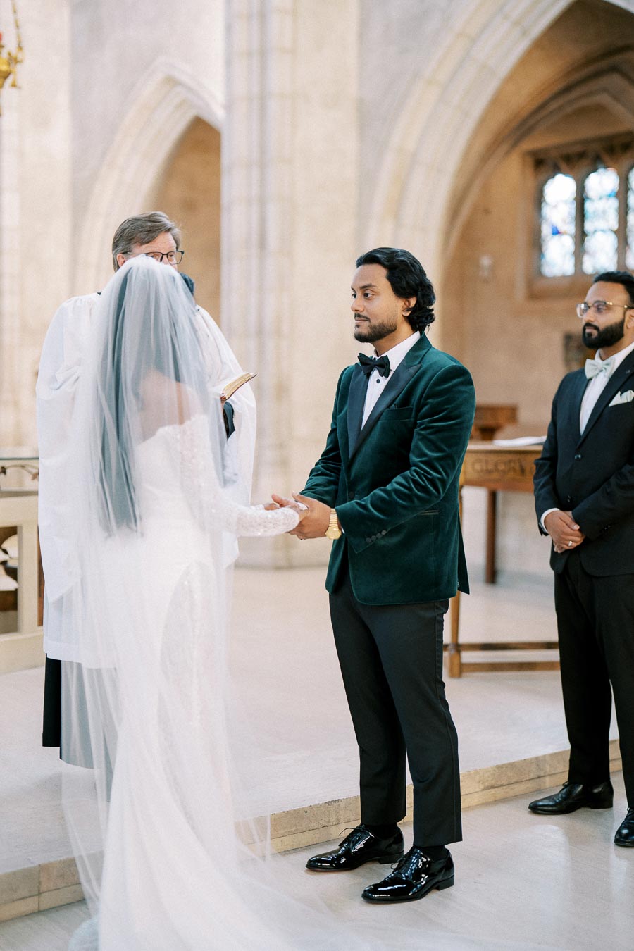 Bride and groom exchanging vows in a church during their wedding ceremony, with the groom wearing a green velvet tuxedo and the officiant reading from a book.