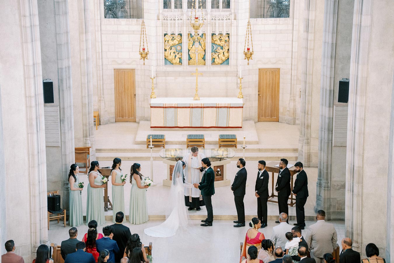 A wedding ceremony taking place in a grand cathedral with a bride and groom standing at the altar, surrounded by bridesmaids in matching dresses and groomsmen in suits, while a priest officiates.