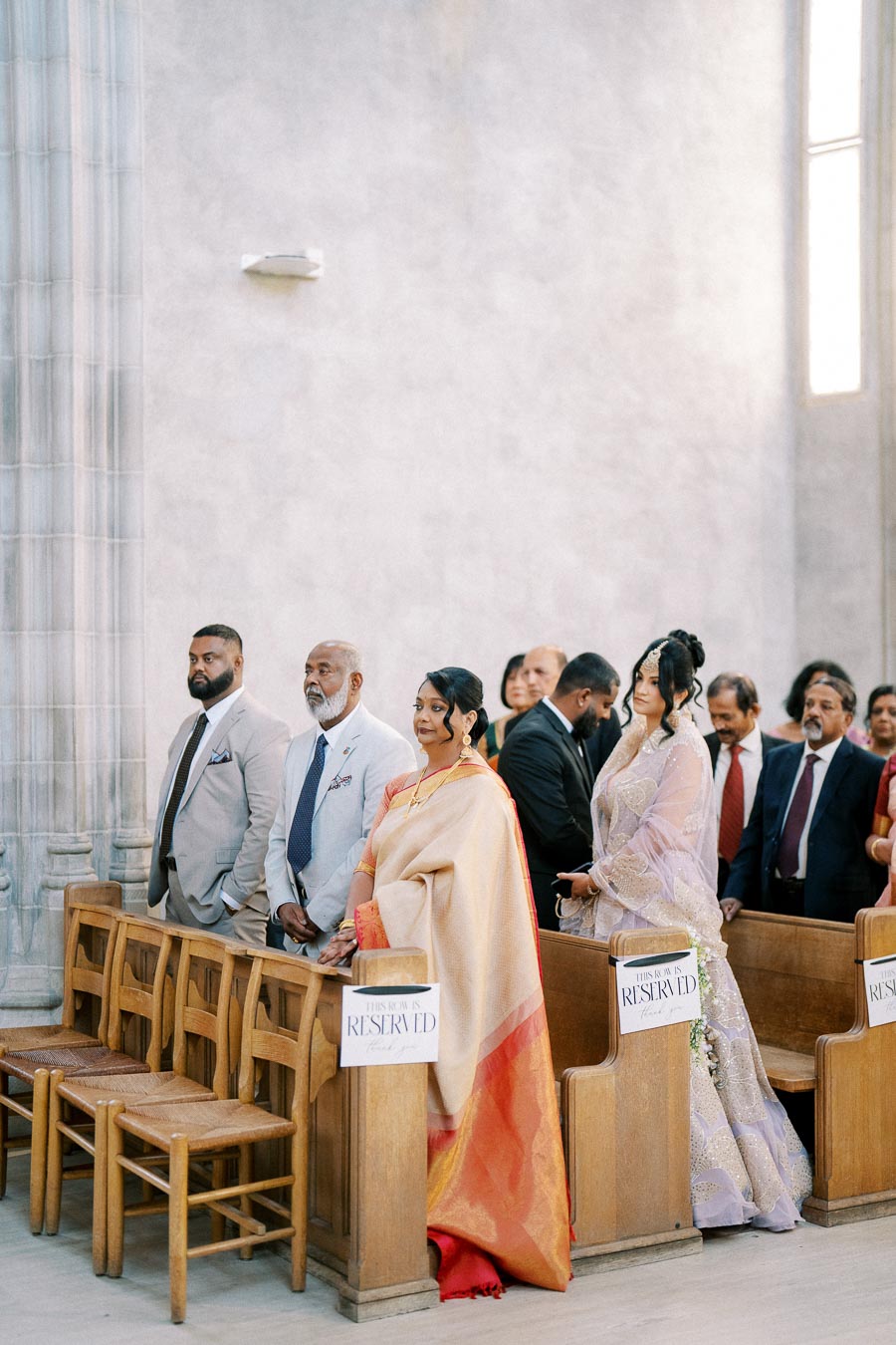 A diverse group of elegantly dressed wedding guests standing in a reserved section inside a church, showcasing a blend of traditional and modern attire.
