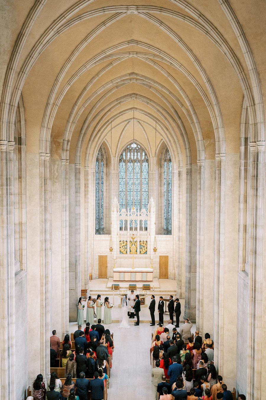 A grand cathedral interior during a wedding ceremony, featuring high arched ceilings, a large stained glass window, and an ornate altar. Guests are seated in pews, while the bridal party stands at the front.