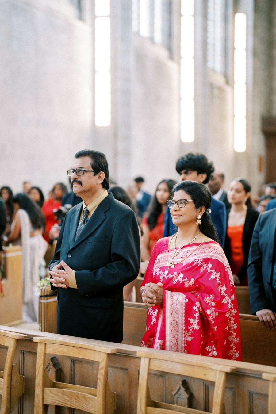 A well-dressed couple stands inside a beautifully lit church during a ceremony, with the woman in a vibrant red saree and the man in a dark suit, surrounded by other attendees.