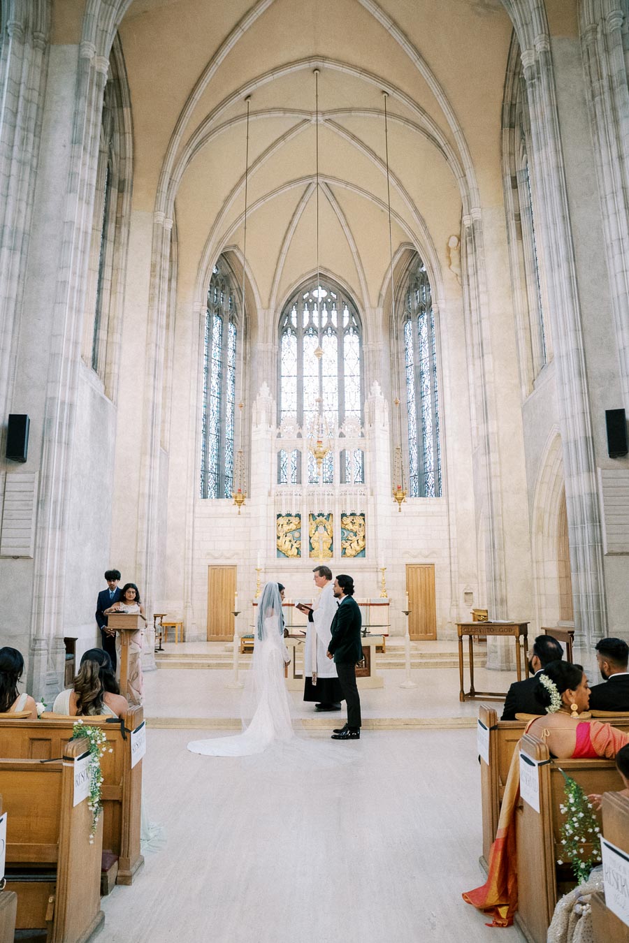 A couple exchanging vows in a large, ornate church with high ceilings and intricate stained glass windows, surrounded by wedding guests seated in pews.