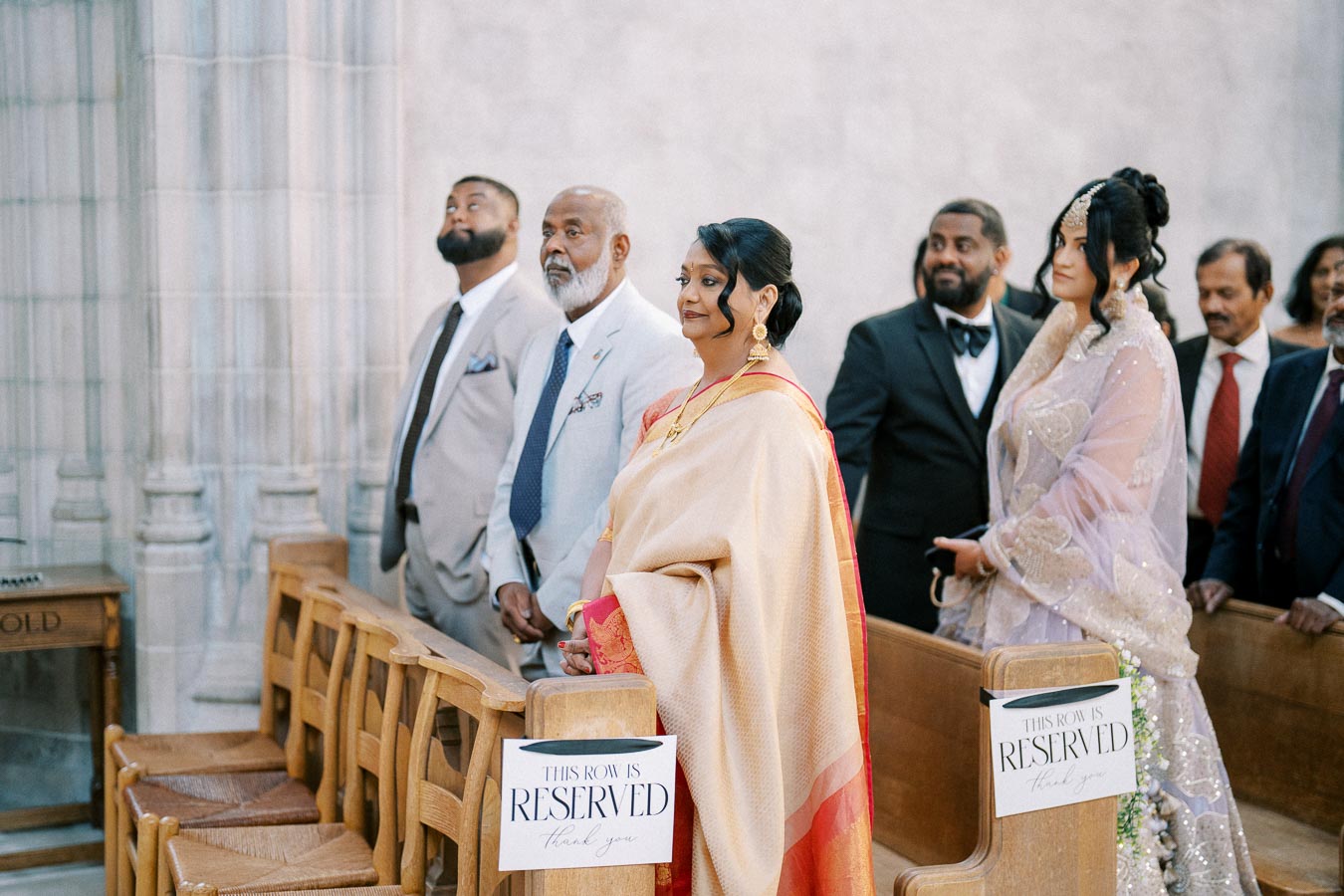 Wedding guests standing in an ornate hall, wearing formal attire including a saree and suits, next to wooden chairs with “Reserved” signs.