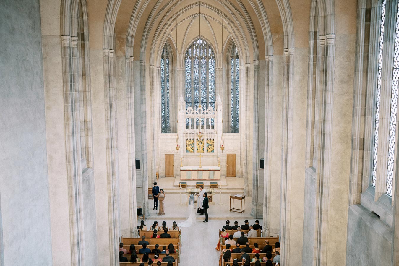 Wedding ceremony inside a grand cathedral with vaulted ceilings, featuring a couple at the altar, surrounded by guests seated in pews.