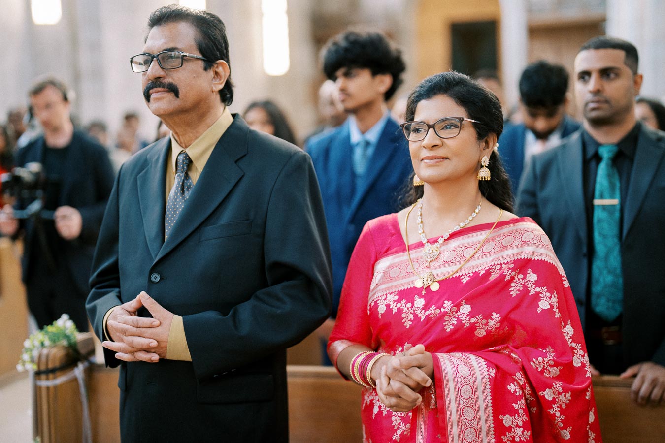 Elegant Indian couple attending a formal event, the woman in a pink saree with gold jewelry and the man in a black suit, surrounded by a diverse group of people in a ceremonial setting.