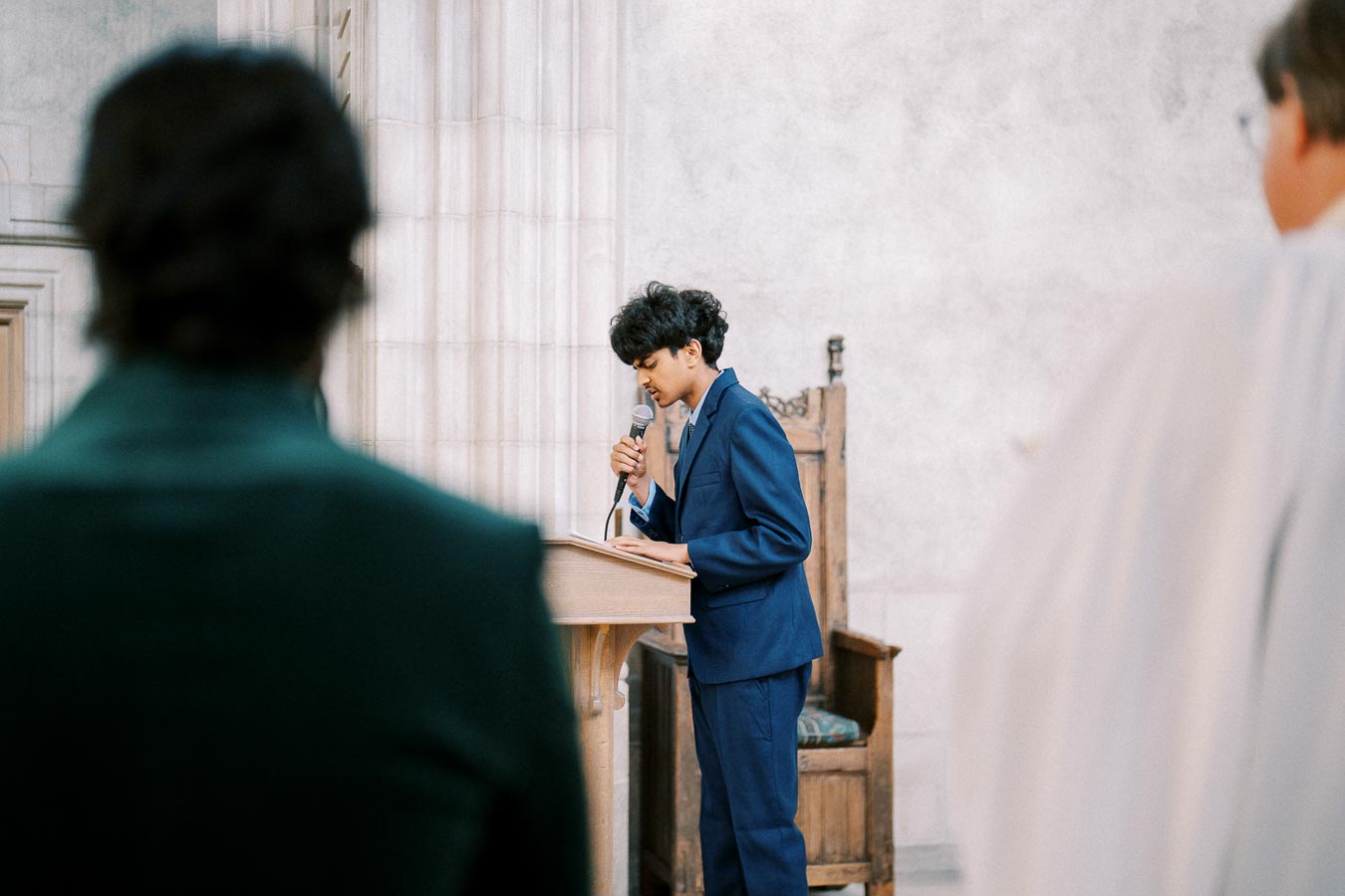 Young speaker in a blue suit presenting at a podium in a formal gathering, with blurred audience members in the foreground.