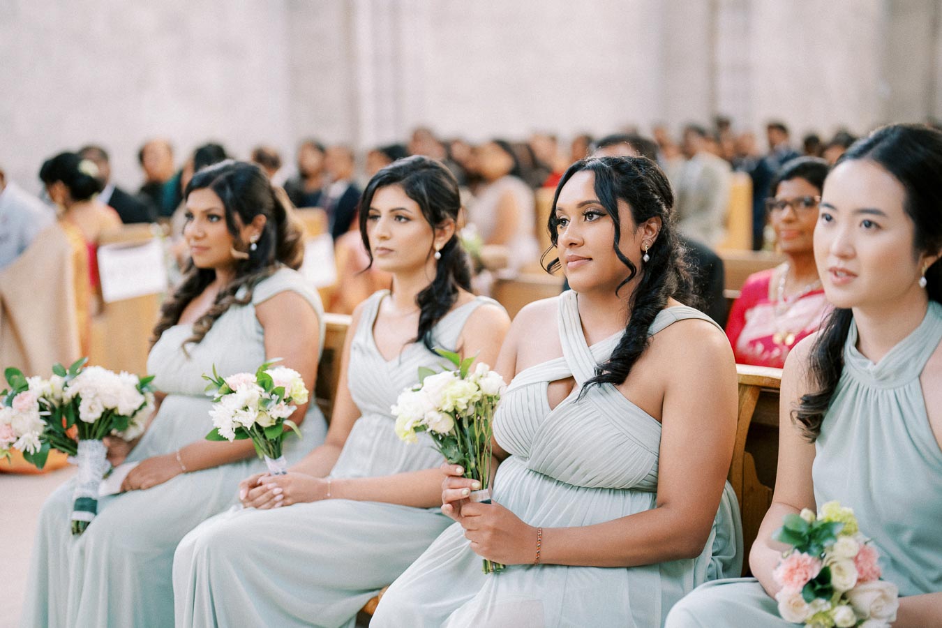 Four bridesmaids in mint green dresses holding bouquets, seated in a church during a wedding ceremony, with a blurred background of other guests.