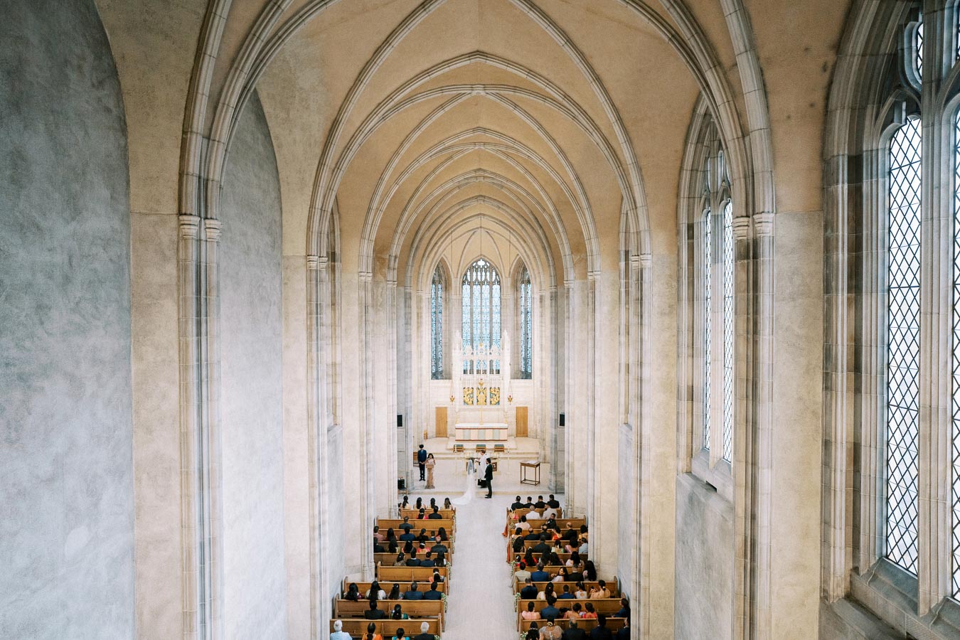 Interior view of a grand cathedral during a wedding ceremony, featuring high vaulted ceilings, stained glass windows, and rows of seated guests in wooden pews.