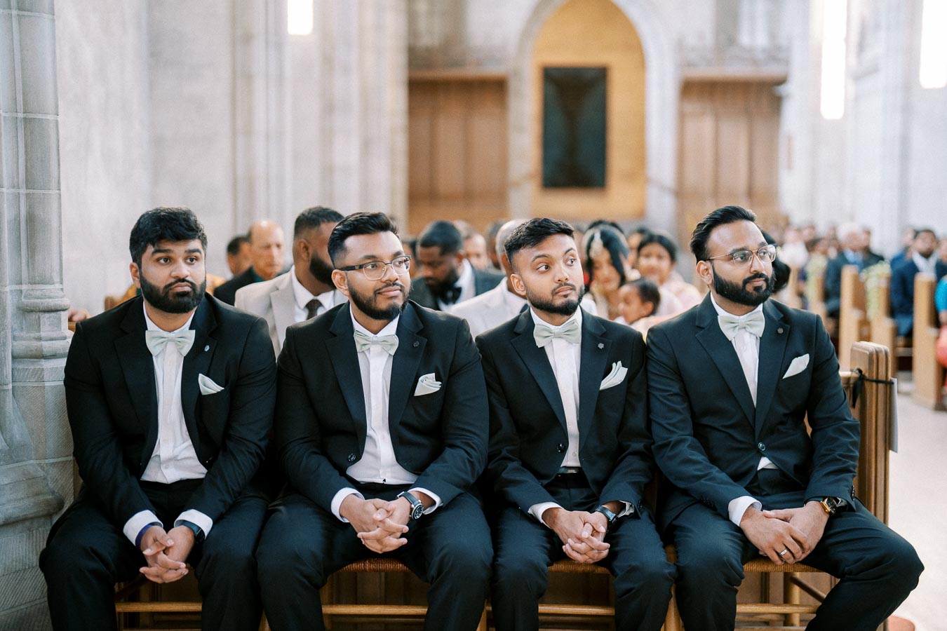 Four groomsmen in matching suits and bow ties seated in a church, waiting at a wedding ceremony.