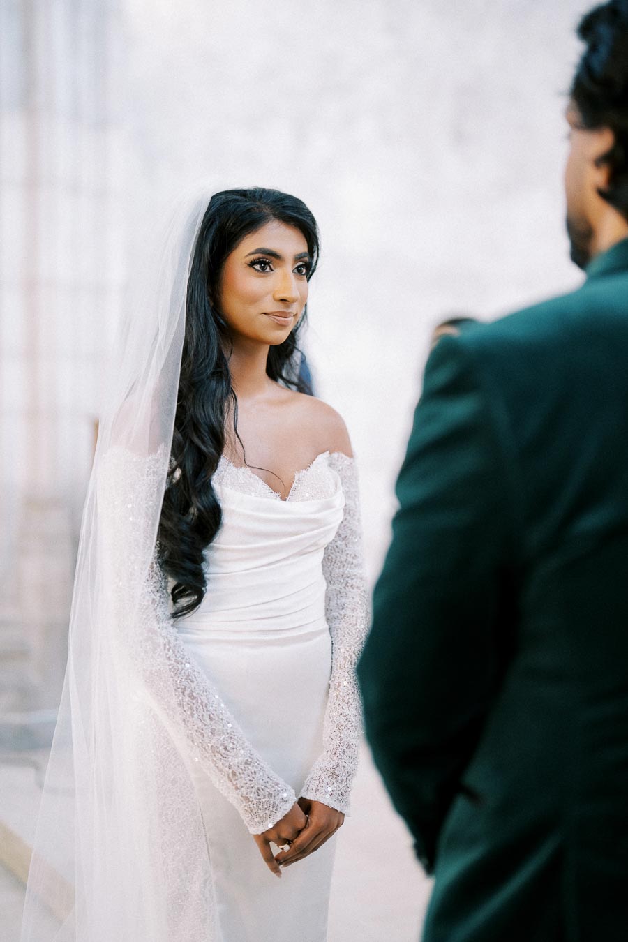 A bride in an elegant white wedding dress and lace sleeves, standing with hands clasped and gazing towards her partner during a wedding ceremony.