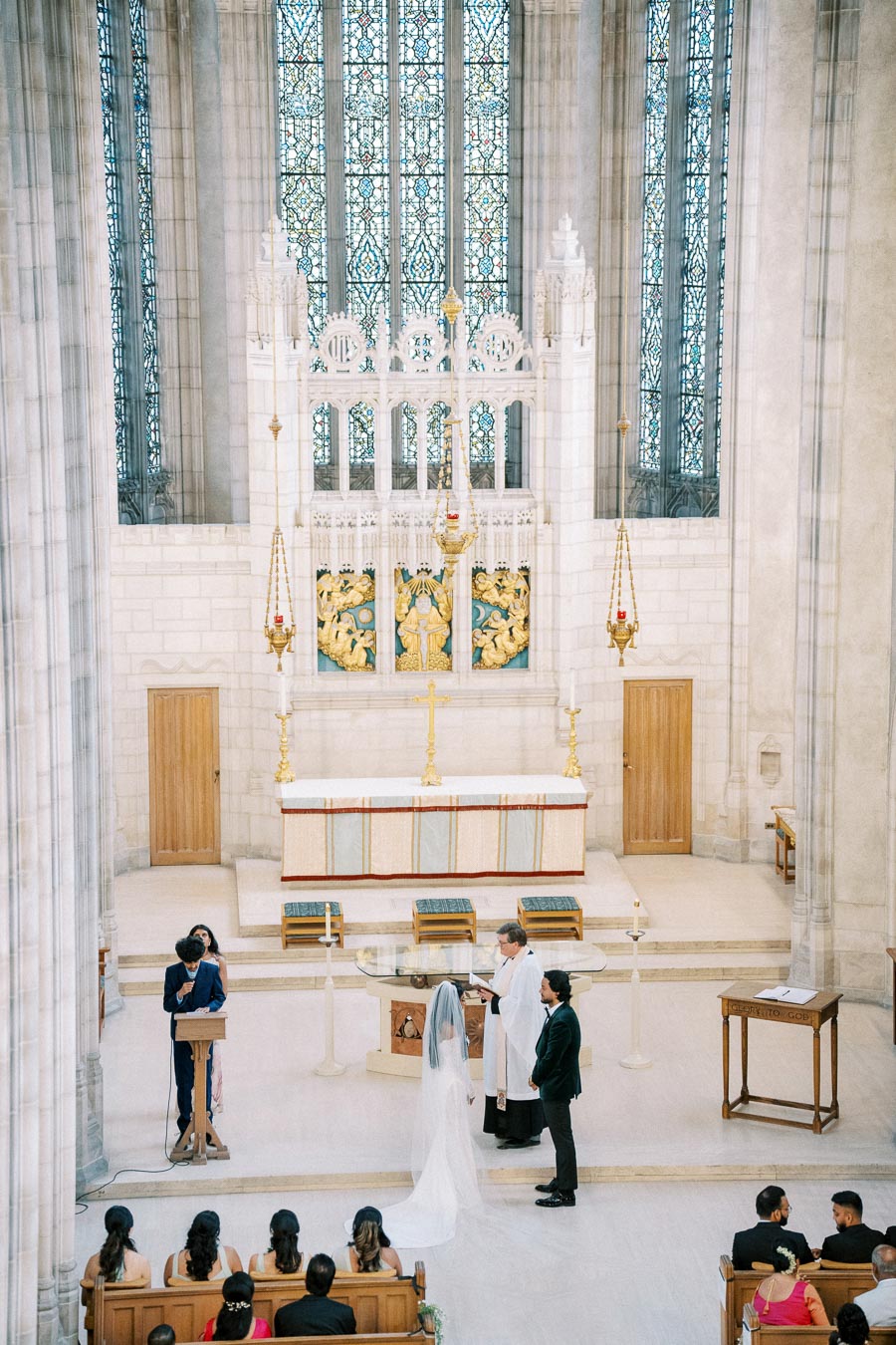 Elegant church wedding ceremony with bride and groom standing at the altar, surrounded by ornate stained glass windows and white stone architecture, while guests look on from wooden pews.
