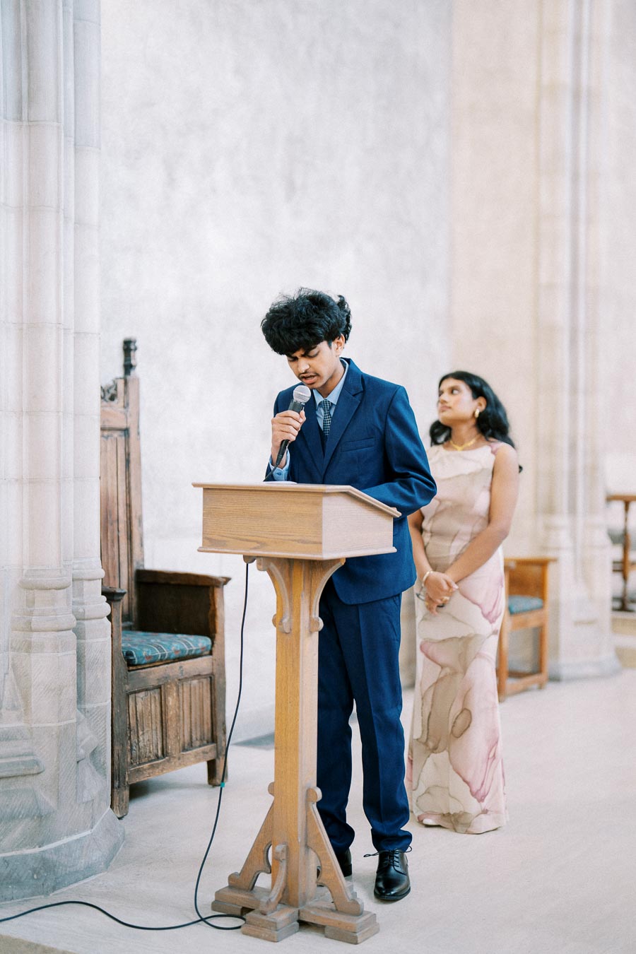 Young man in a blue suit speaking at a podium with a microphone, while a young woman in a floral dress stands attentively behind him in a formal setting.