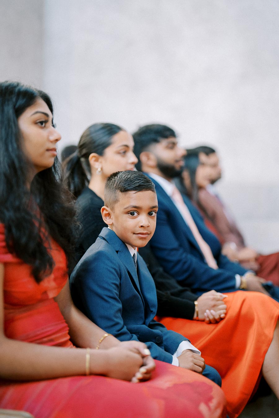 Group of people, including a young boy in a blue suit, sitting attentively during a formal event, dressed in elegant attire.