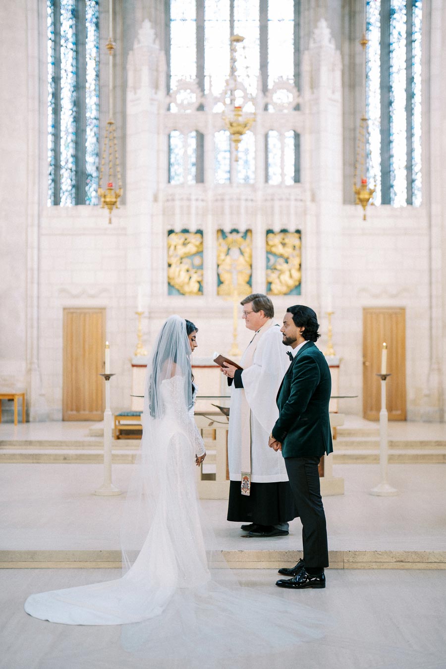 A bride and groom stand before a priest during their wedding ceremony in a beautifully adorned church, with intricate stained glass windows and elegant decor.