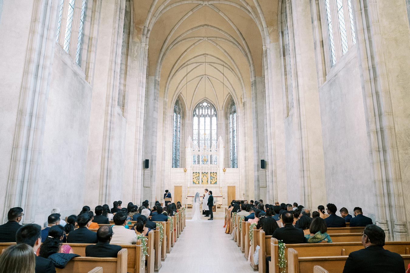A wedding ceremony taking place in a grand, gothic-style church with high vaulted ceilings and large stained glass windows, attended by a seated congregation.