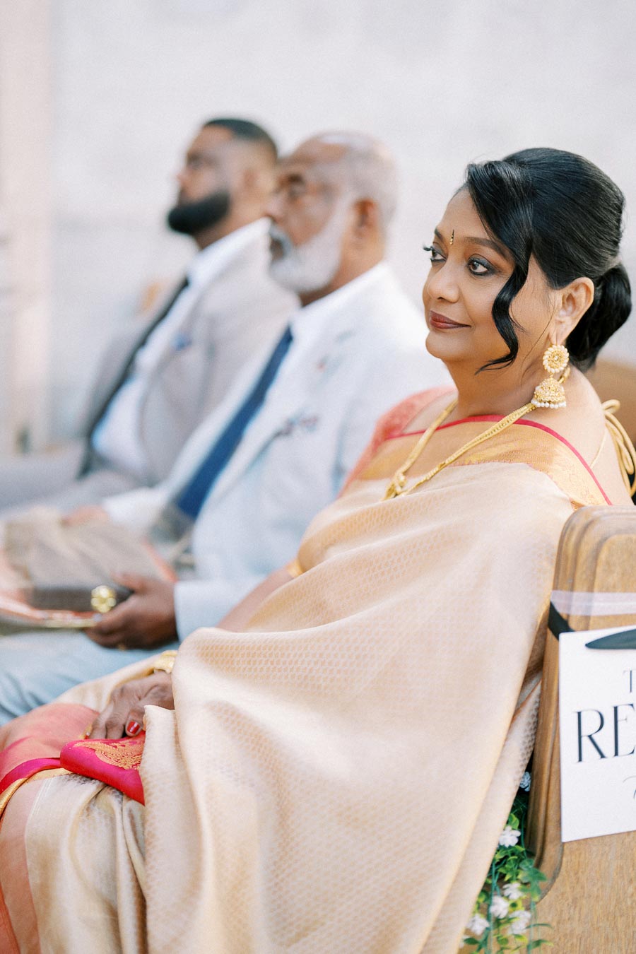 A woman in traditional attire sits pensively during a formal event, adorned with elegant jewelry. Two men in suits sit nearby, all appearing attentive and engaged.