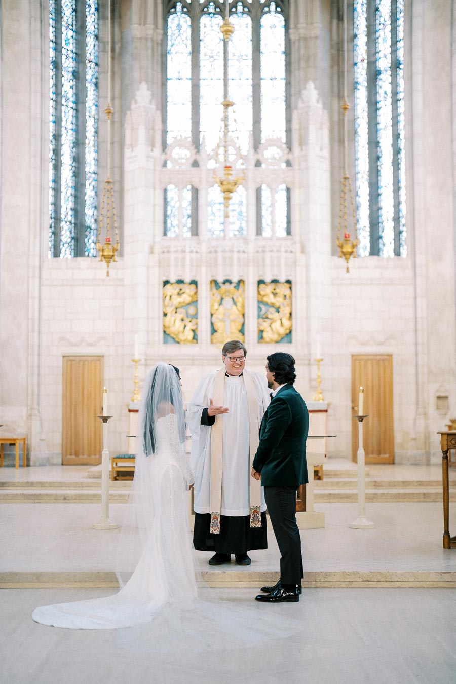 A bride and groom stand before an officiant in a grand church setting, with elegant stained glass windows and intricate decorations, during a wedding ceremony.