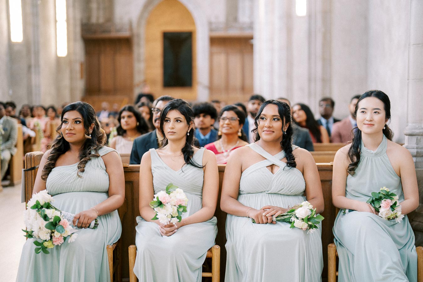 Four bridesmaids in matching light grey dresses sitting in a church pew, holding bouquets of pink and white flowers, with a wedding ceremony in progress in the background.