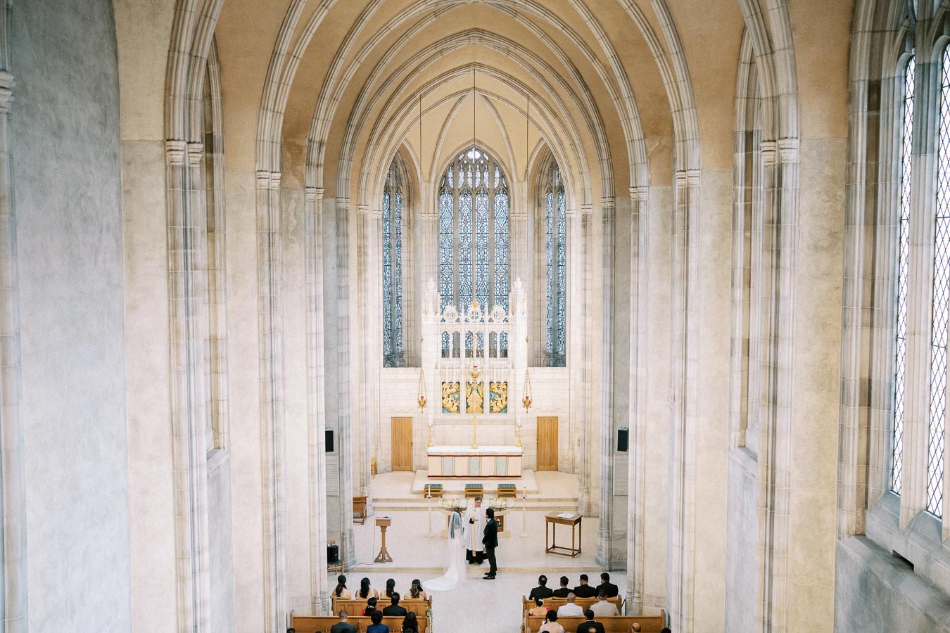 Aerial view of a couple getting married in a grand cathedral with vaulted ceilings and large stained glass windows, surrounded by seated guests.