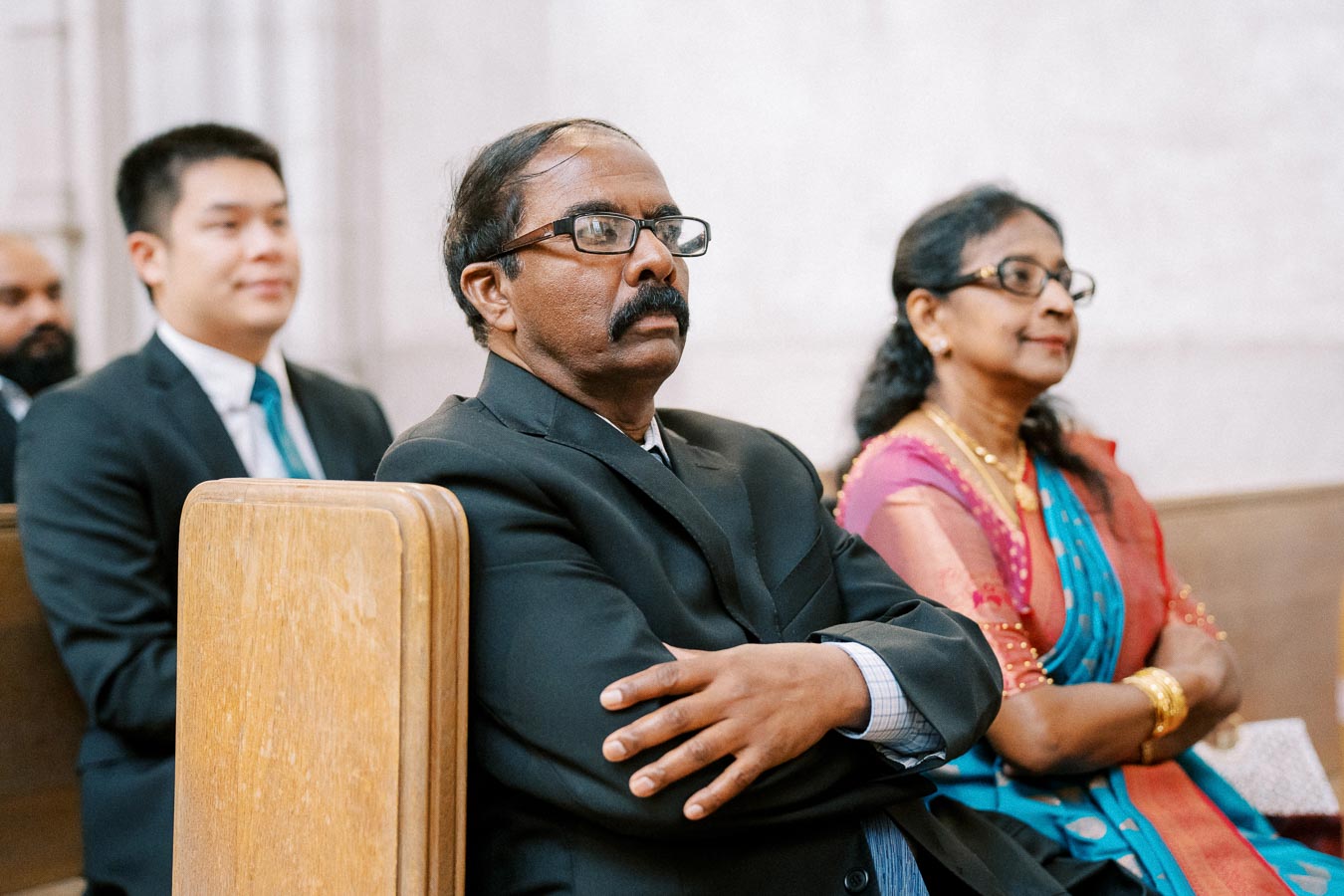A group of people seated attentively in a church pew during a formal event, including a man in a suit and tie and a woman in traditional attire.