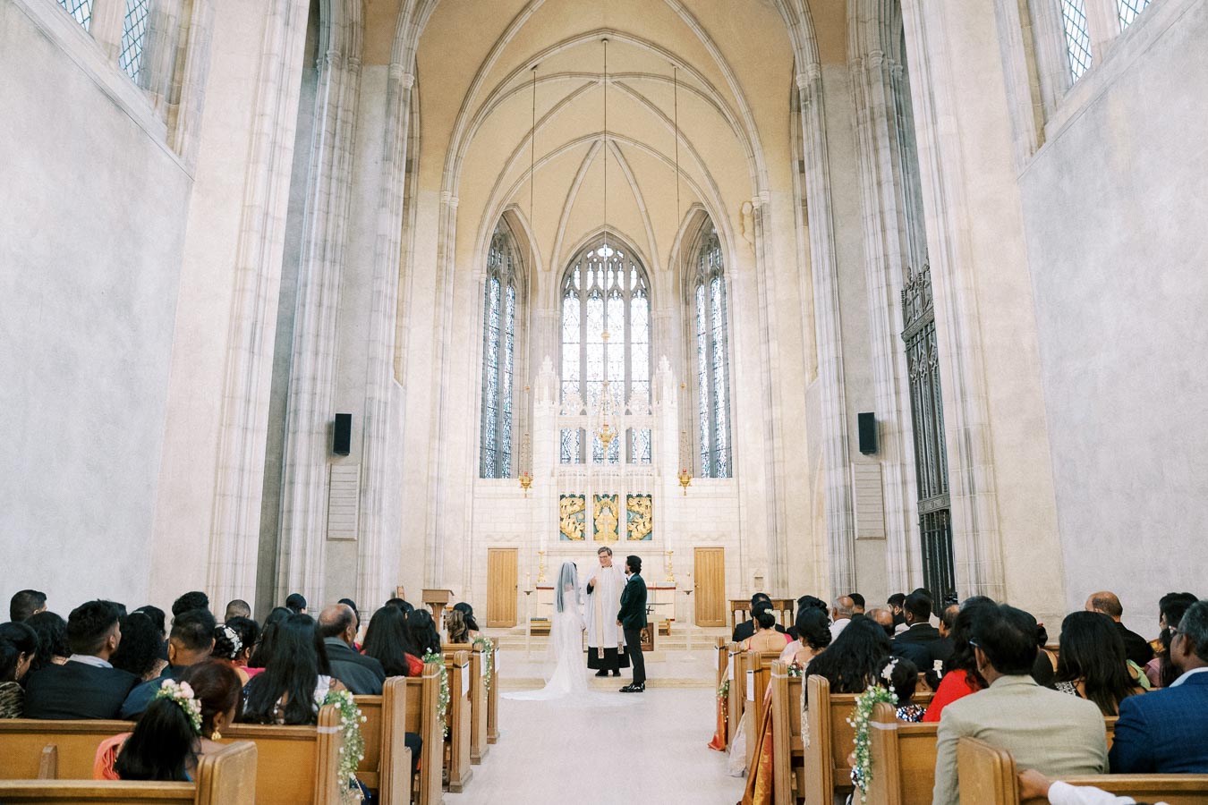 Elegant wedding ceremony inside a grand cathedral with high vaulted ceilings and large stained-glass windows, with the couple standing at the altar and guests seated in pews.