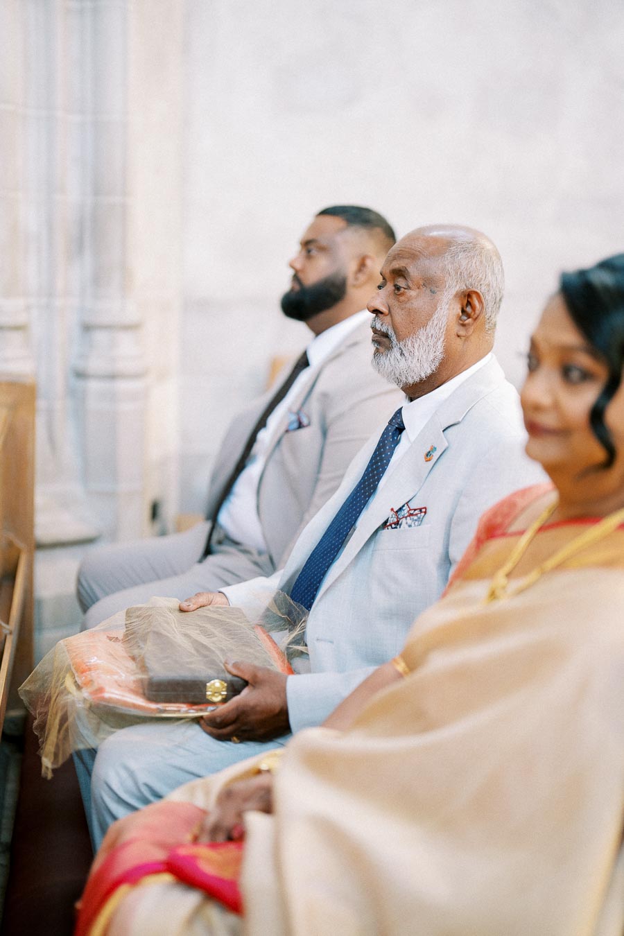 A group of elegantly dressed people seated in a church, with one man in a light-colored suit holding wrapped gifts, possibly attending a wedding ceremony.