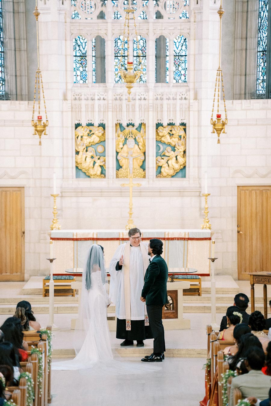 A bride and groom stand before a priest in a beautifully decorated church during their wedding ceremony, surrounded by guests.