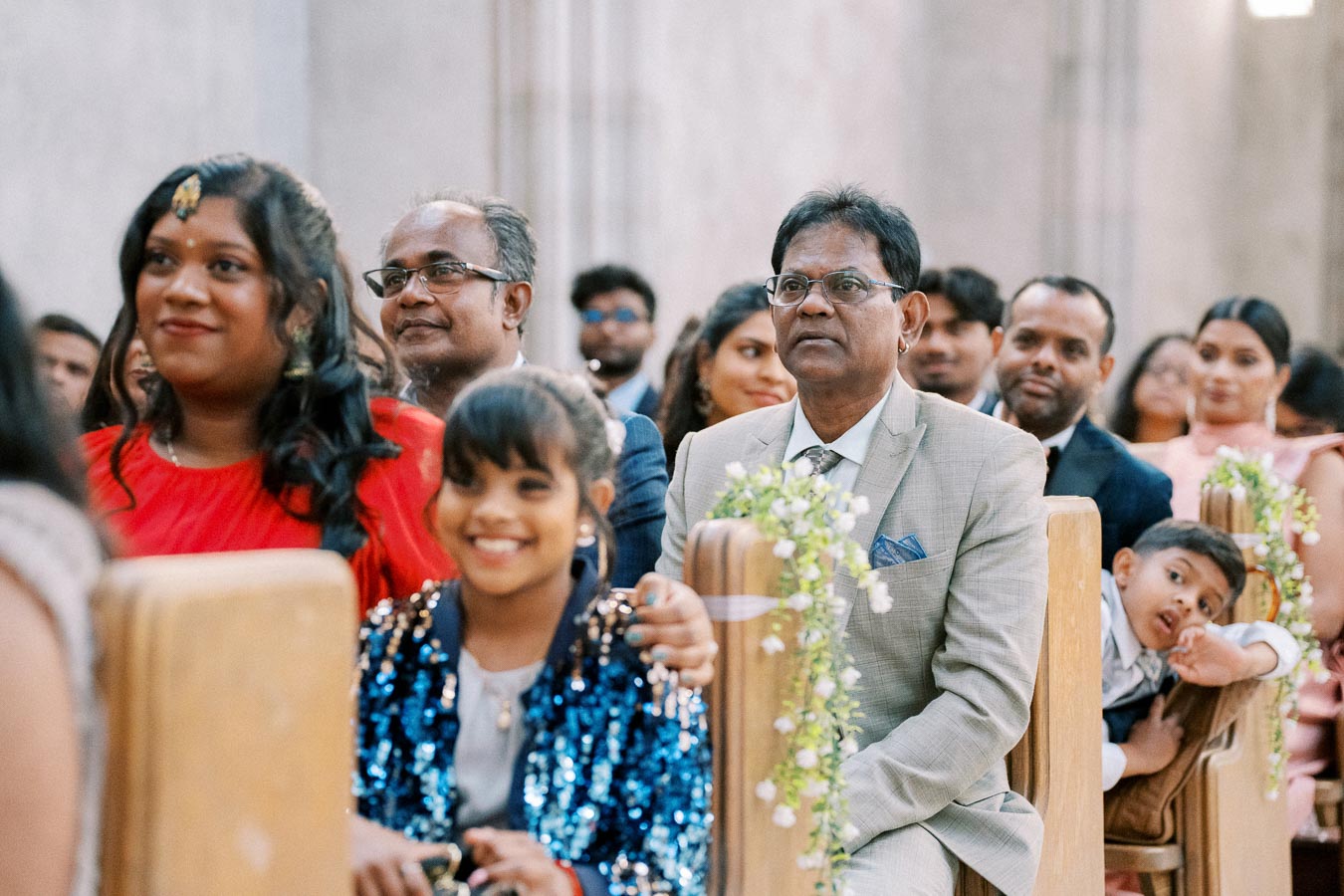 A group of people seated in a well-decorated indoor venue, attentively watching an event. The setting appears formal, with individuals dressed in elegant attire, suggesting a special occasion or ceremony.