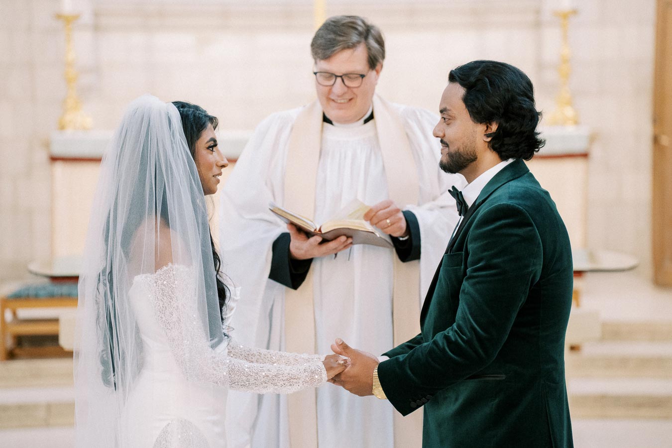 A bride and groom hold hands and exchange vows in a church wedding ceremony, with a priest officiating in the background. The bride wears a strapless white gown and veil, while the groom is dressed in a dark green velvet suit.