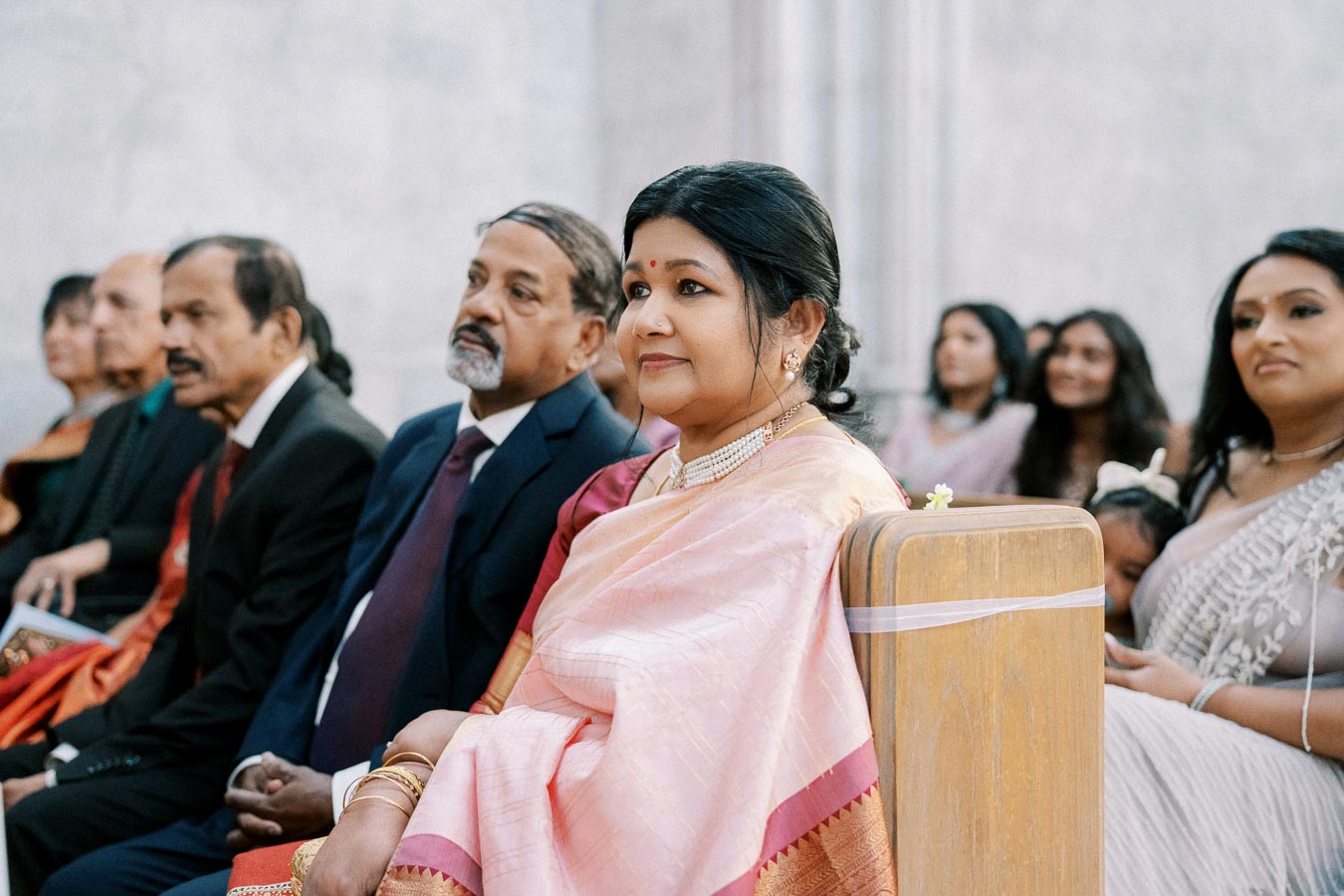Group of people seated in a formal setting, with a focus on a woman wearing a pink sari and gold jewelry, attending what appears to be a ceremonial event.