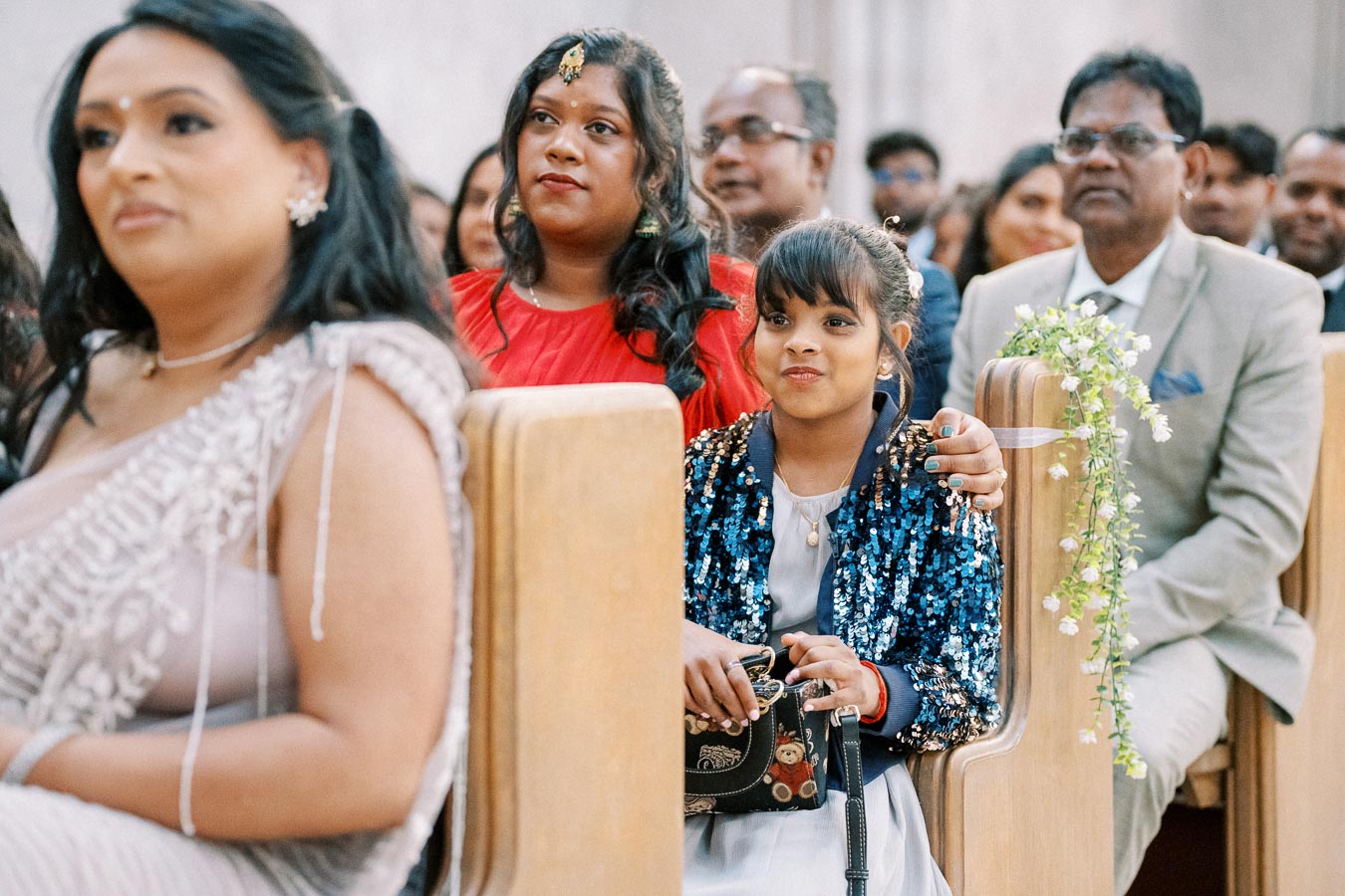 A diverse group of people sitting attentively in wooden pews during a formal event, including a young girl in a blue sequined jacket and a woman in a red dress; floral decorations are visible on the pews.