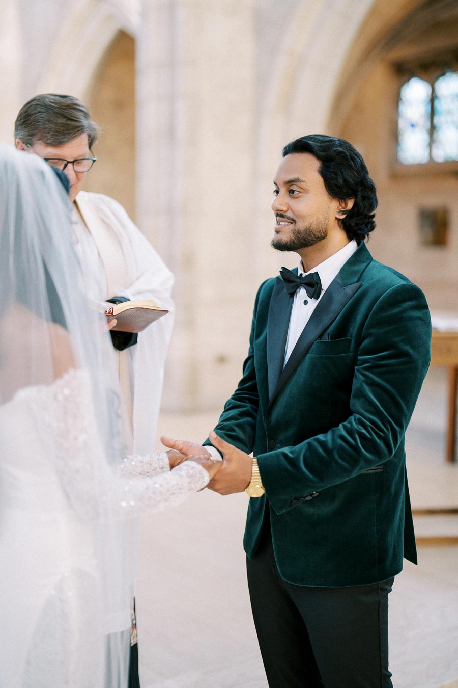 Bride and groom exchanging vows in a church ceremony, groom in a green velvet tuxedo holding the bride's hands, with a priest officiating the wedding.
