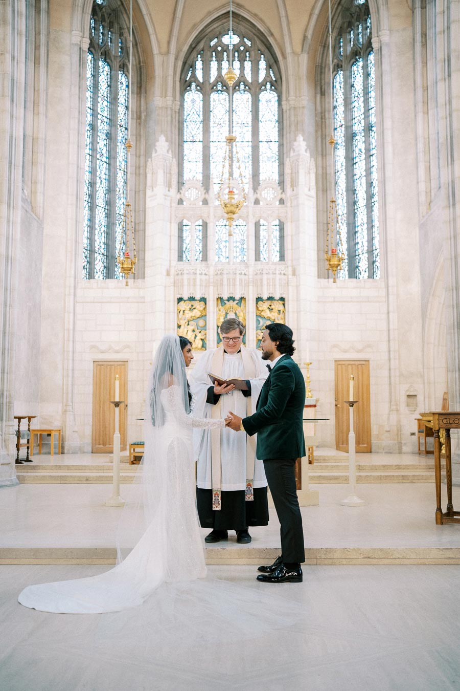 A couple exchanges vows during a wedding ceremony in a grand church with beautiful stained glass windows, presided over by a minister in traditional attire.