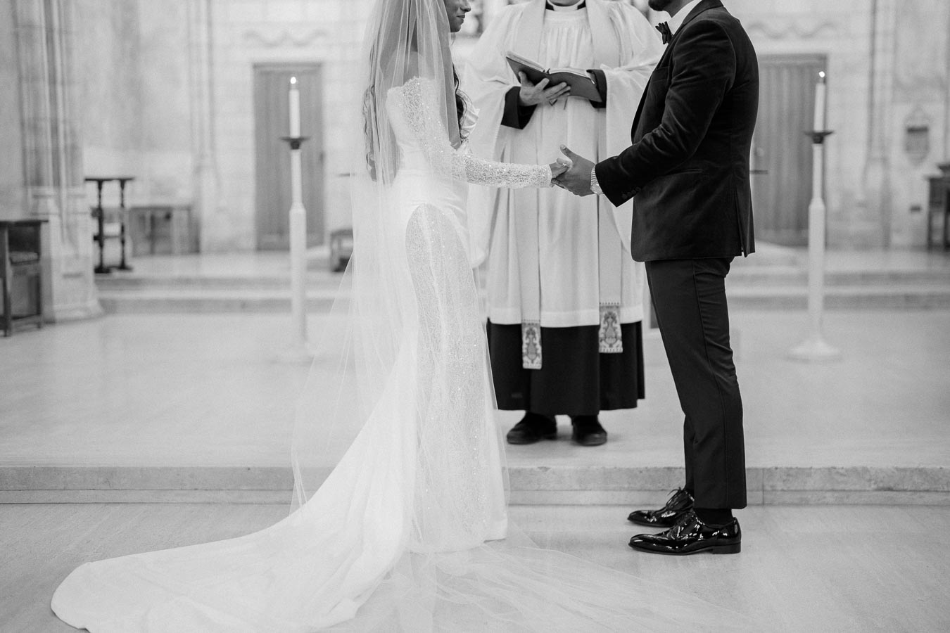 Black and white image of a wedding ceremony with a bride in a long gown and veil holding hands with the groom, while an officiant in robes conducts the ceremony in an elegant church setting.