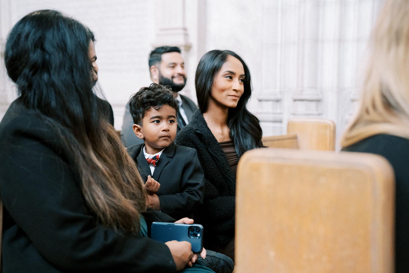 A group of people dressed formally, including a young boy in a suit, sit attentively on wooden benches inside a bright, ornate room, suggesting a formal event or ceremony.