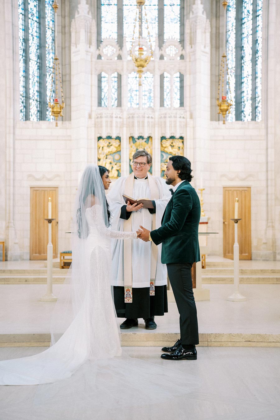 Bride and groom holding hands during a wedding ceremony in a cathedral, with a priest officiating, surrounded by ornate architecture and candles.